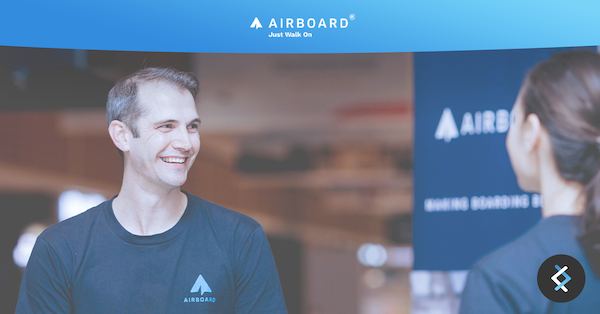 photo of man smiling wearing Airboard tshirt, looking at a woman. Airboard sign in the background. Above is white airboard logo on a blue banner background
